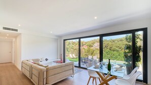 A modern living room with a sofa, a glass dining table, and chairs. There is a large window offering a view of a garden and buildings. - Villa in Lloret de Mar with Private Pool (Vidreres)