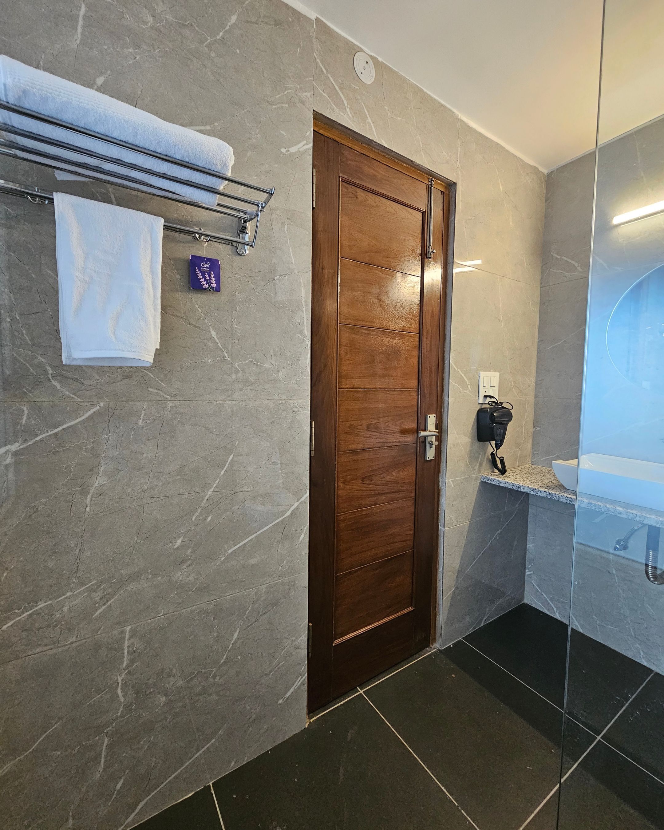 A modern bathroom with a wooden door, marble walls, a glass shower enclosure, and a towel rack with folded towels.