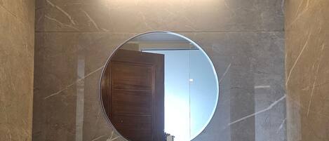Un baño con un espejo redondo, un lavamanos blanco y una lámpara empotrada en la pared.