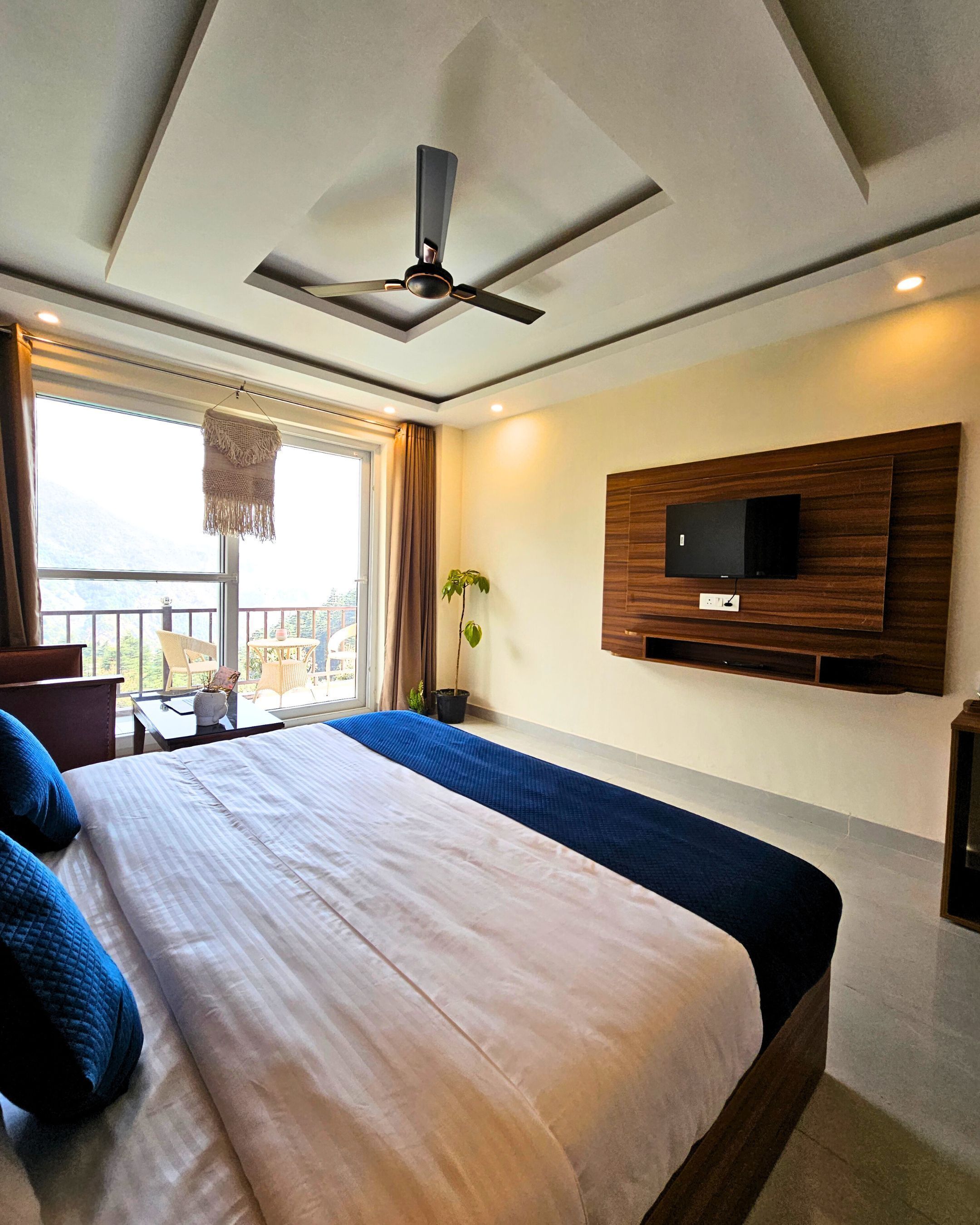 A modern hotel room with a large bed, a ceiling fan, a TV mounted on the wall, and a balcony with a view.
