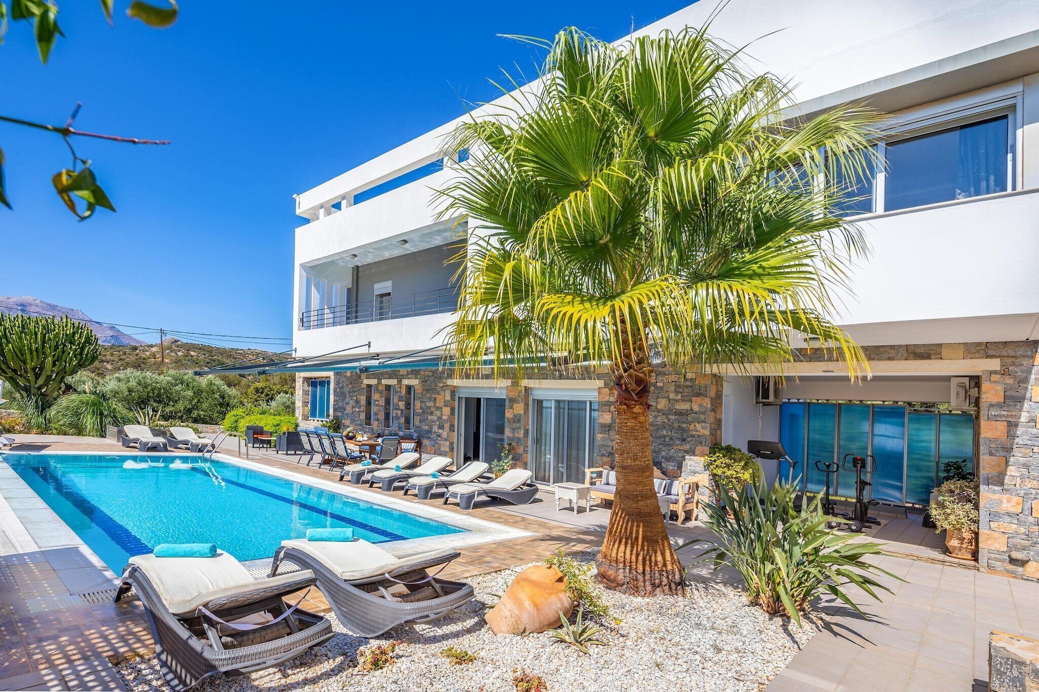 Un bâtiment moderne à deux étages, doté d’une piscine, de chaises longues et d’un palmier.