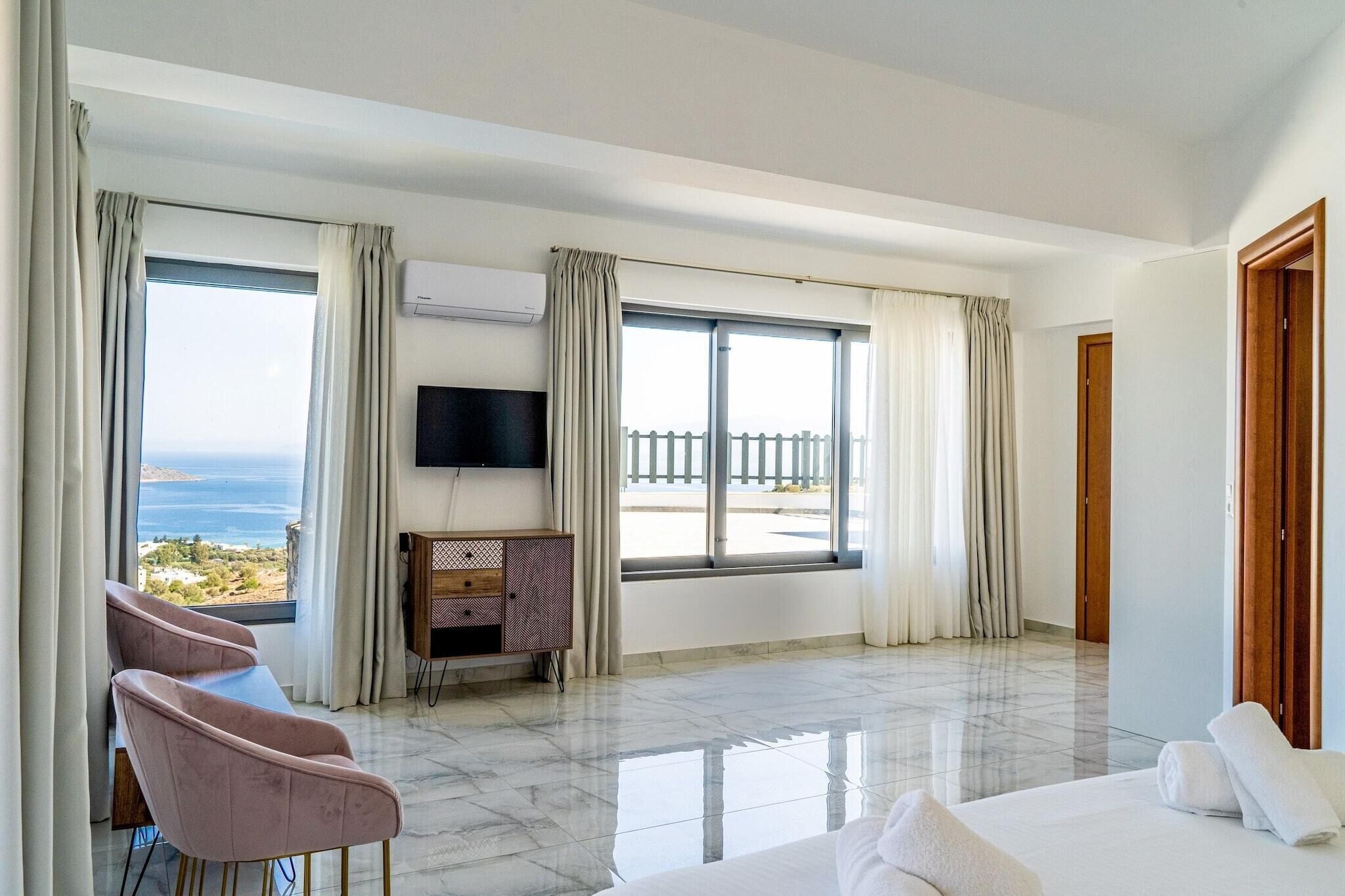 Una habitación de hotel moderna con una cama grande, una zona de estar con sillones, un televisor montado en la pared y un balcón con vistas al mar.