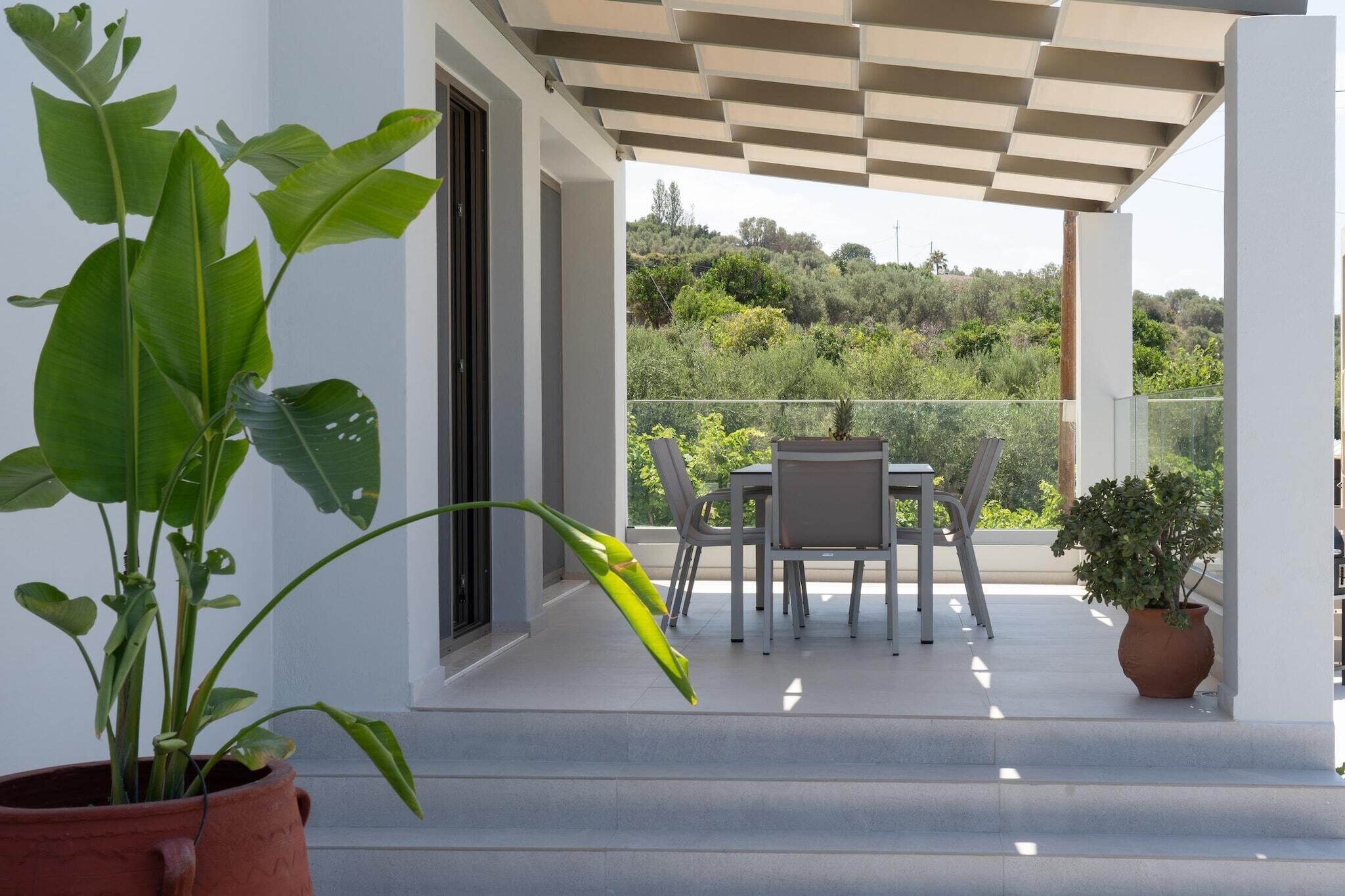 Un balcón con una mesa y sillas, una planta en maceta, y vistas de follaje al fondo.