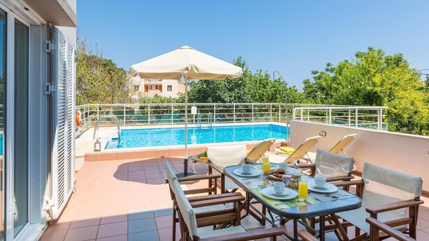 A rooftop pool area with a table set for breakfast, a lounge chair, and a view of a building in the background.