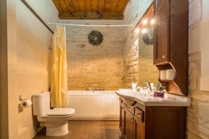 Un bagno con parete in pietra, soffitto in legno, vasca, water e mobile lavabo con specchio.
