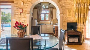 A stone-walled room with a glass dining table, chairs, a TV, and a kitchen visible through an archway. - Villa in Rethymno with countryside view (Rethymno)