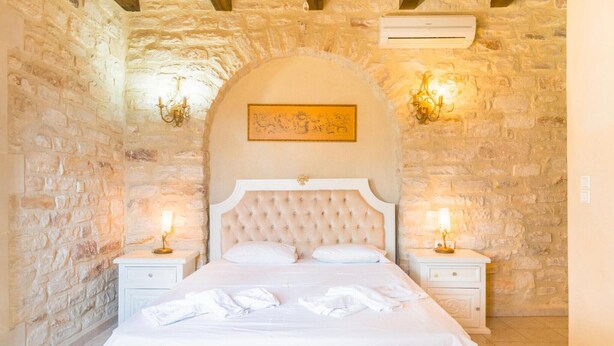 A bedroom with a stone wall, a tufted headboard, two bedside tables with lamps, and a bed with white linens. - Villa in Rethymno with countryside view (Rethymno)