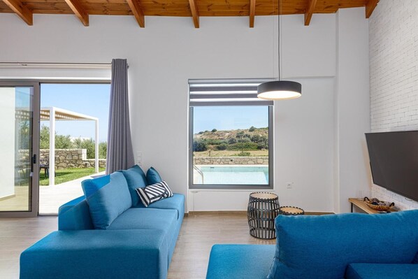 Una sala de estar moderna con un sofá azul, un ventanal con vistas a una piscina y un paisaje, y un televisor de pantalla plana montado en la pared.