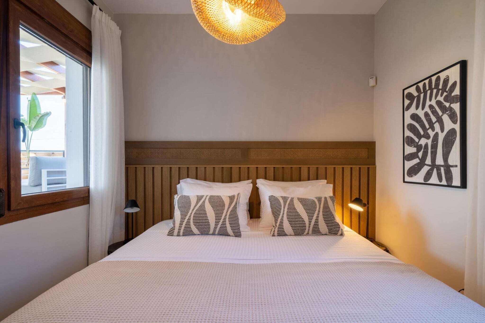 Une chambre d’hôtel avec un lit, deux coussins à motifs, un luminaire suspendu, une œuvre d’art accrochée au mur et une fenêtre avec des rideaux.