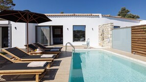 A modern outdoor pool area with lounge chairs and an umbrella, a stone wall, and a white building with large windows. - Scenic villa in Rethymno with private pool (Rethymno)