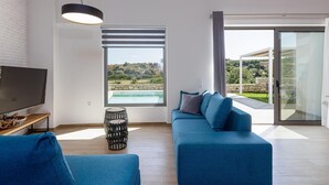 A modern living room with a large TV, a blue sofa, a black hanging light, and a view of a pool and landscape through a window. - Scenic villa in Rethymno with private pool (Rethymno)
