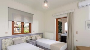 A hotel room with two beds, a window with a blind, a radiator, a small table with a lamp, and a door leading to a bathroom. - Villa in Rethymno with Pool & Jacuzzi (Rethymno)