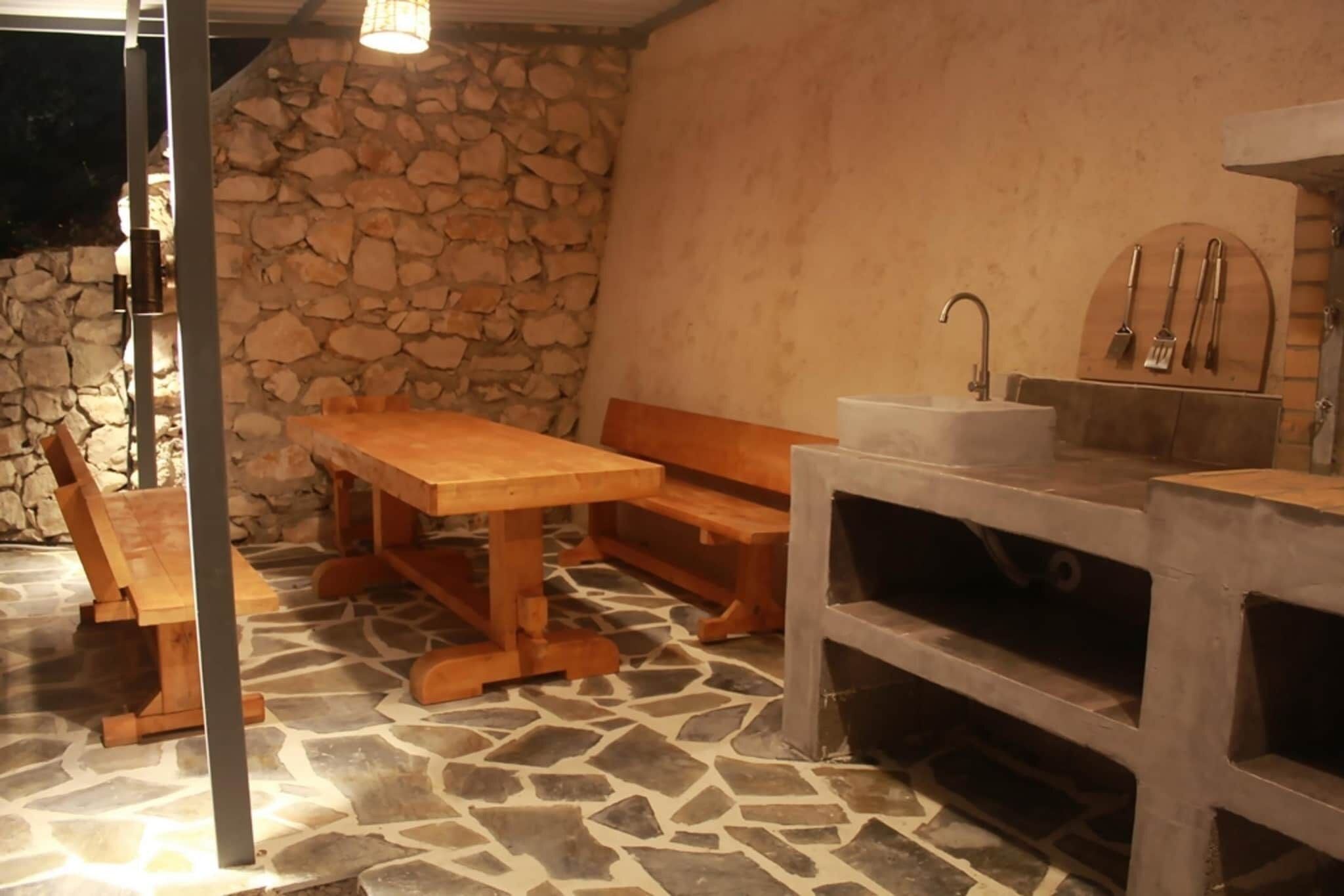 Een buitenkeuken met stenen muren, een houten tafel en banken, een spoelbak en een vloer van natuursteen.