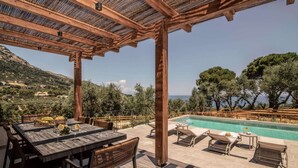 En træpergola over et poolområde med et bord og stole, omgivet af et naturskønt udsigt over træer og bjerge.