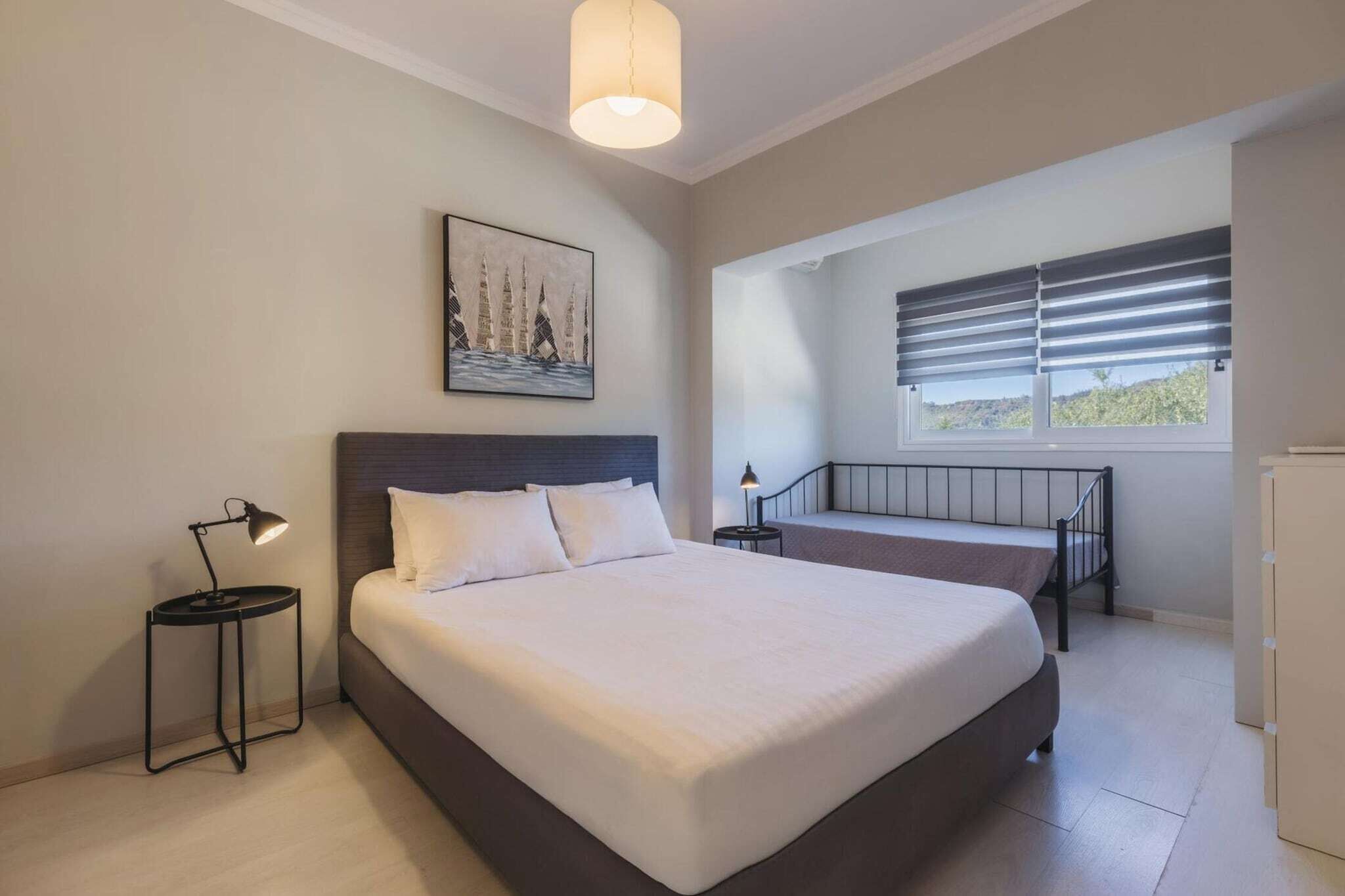 A bedroom with a large bed, a bedside table with a lamp, a window with blinds, and a painting on the wall.