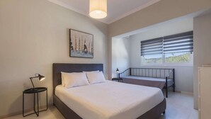 A bedroom with a large bed, a bedside table with a lamp, a window with blinds, and a painting on the wall. - Villa in Kypseli with Private Pool & Gardens (Kipseli)