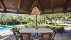 A covered outdoor dining area with a round table and four chairs, a hanging light fixture, and a view of a swimming pool and greenery. - Villa in Kypseli with Private Pool & Gardens (Kipseli)