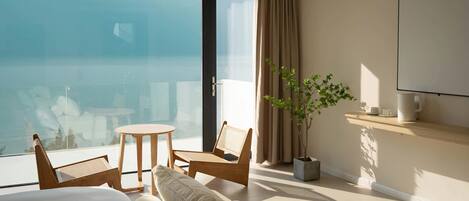 Habitación de hotel con balcón, una cama, un sofá, una mesita, dos sillas, una planta en maceta y un televisor de pared.