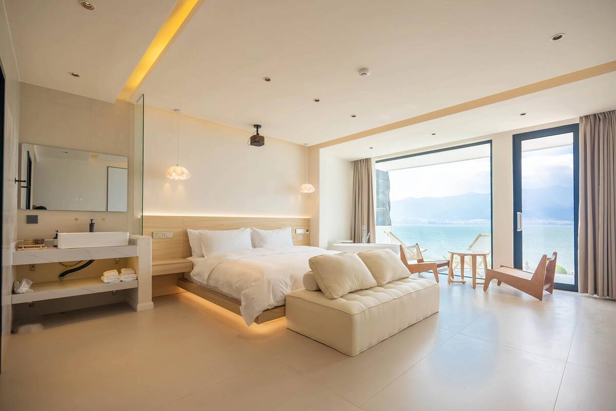 A modern hotel room with a large bed, a sofa, a desk, and a view of the sea.