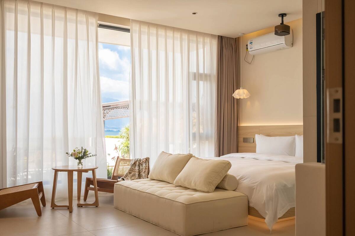 A hotel room with a bed, a sofa, a small table with a vase of flowers, and a view of the outdoors.