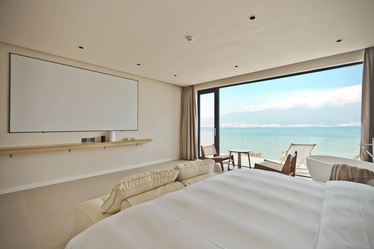 A modern hotel room with a large bed, a flat-screen TV, a small table, and a view of the sea.