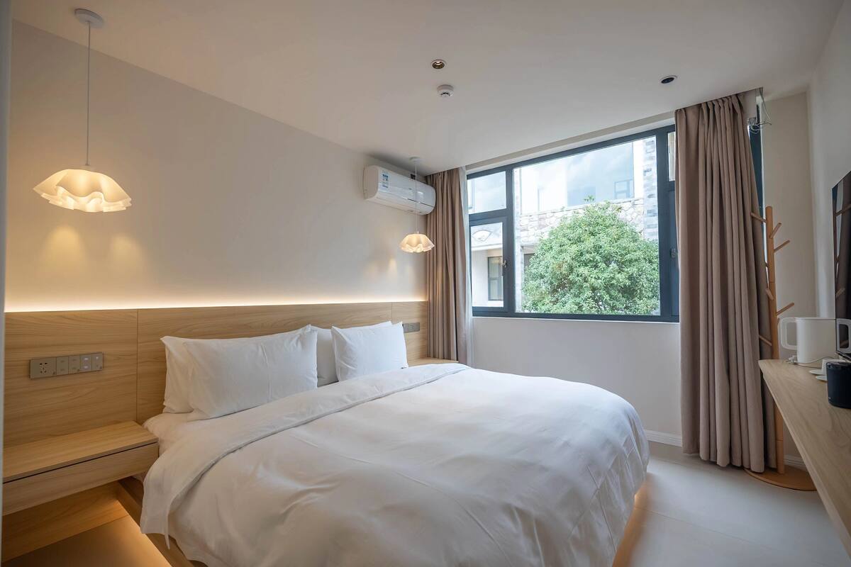 A hotel room with a large bed, bedside tables, a desk with a chair, a window with curtains, and a hanging light fixture.