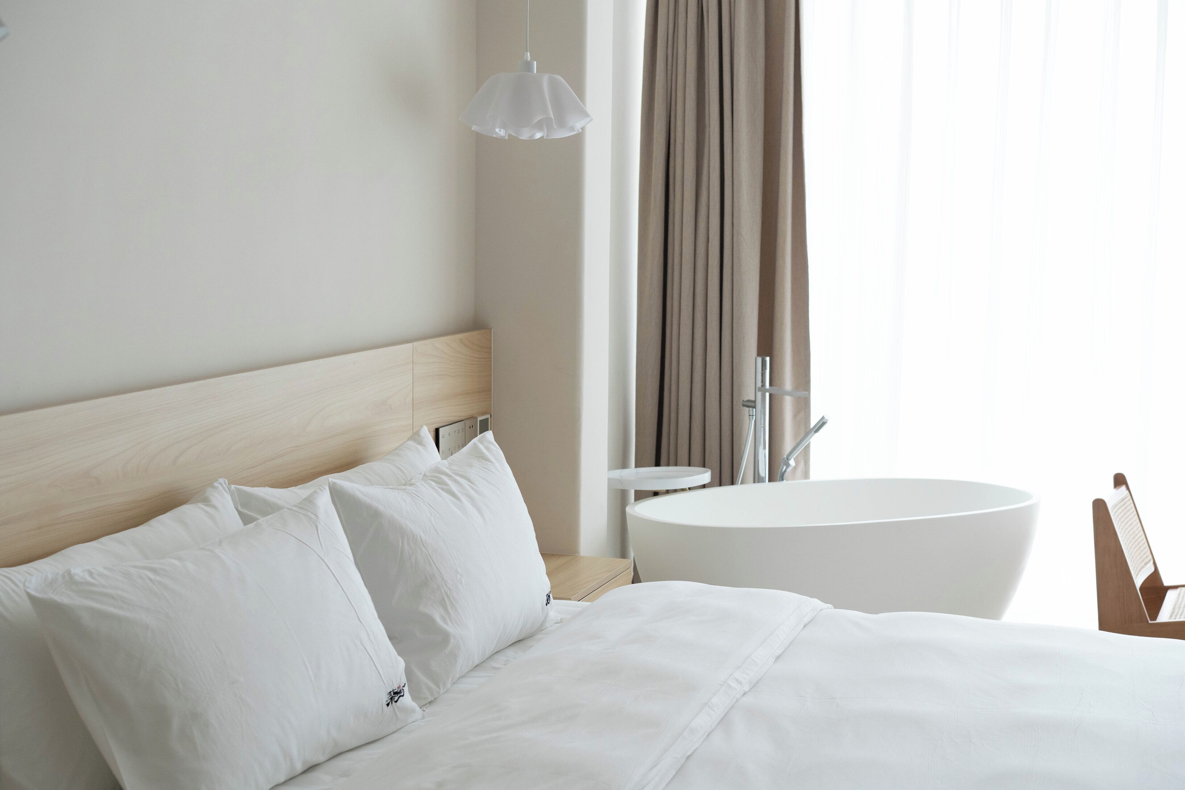 A hotel room with a wooden headboard, a round white bathtub, a white bed with pillows, and a hanging light fixture.