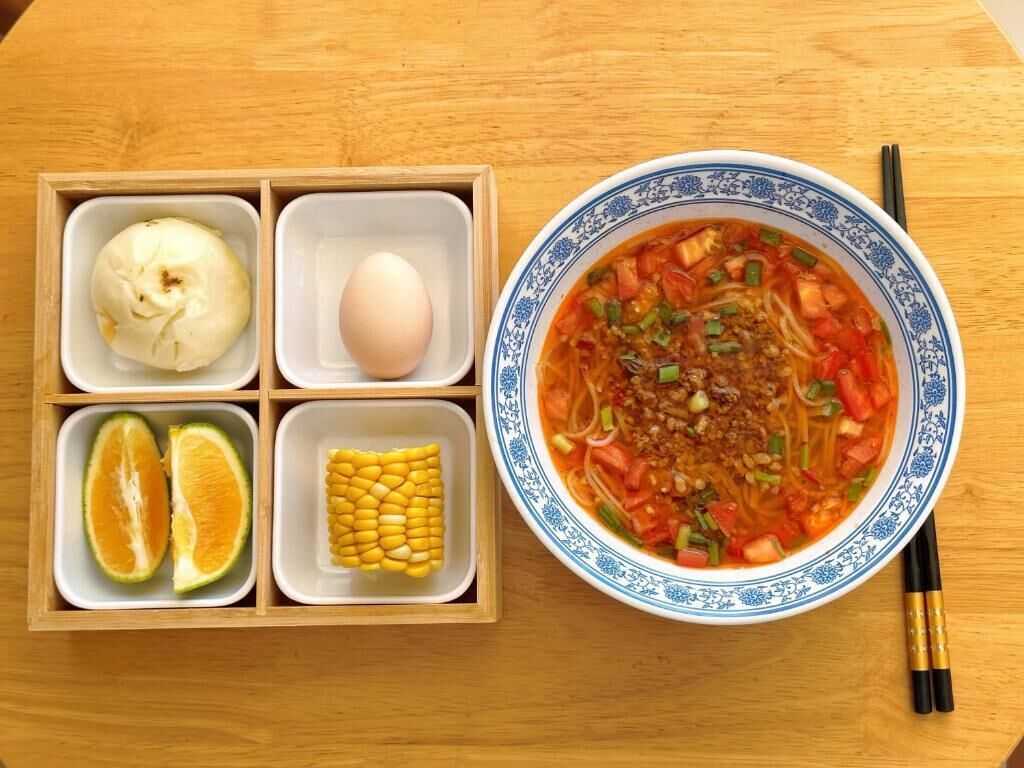 A wooden table with a bowl of soup, a box of food items, and chopsticks.