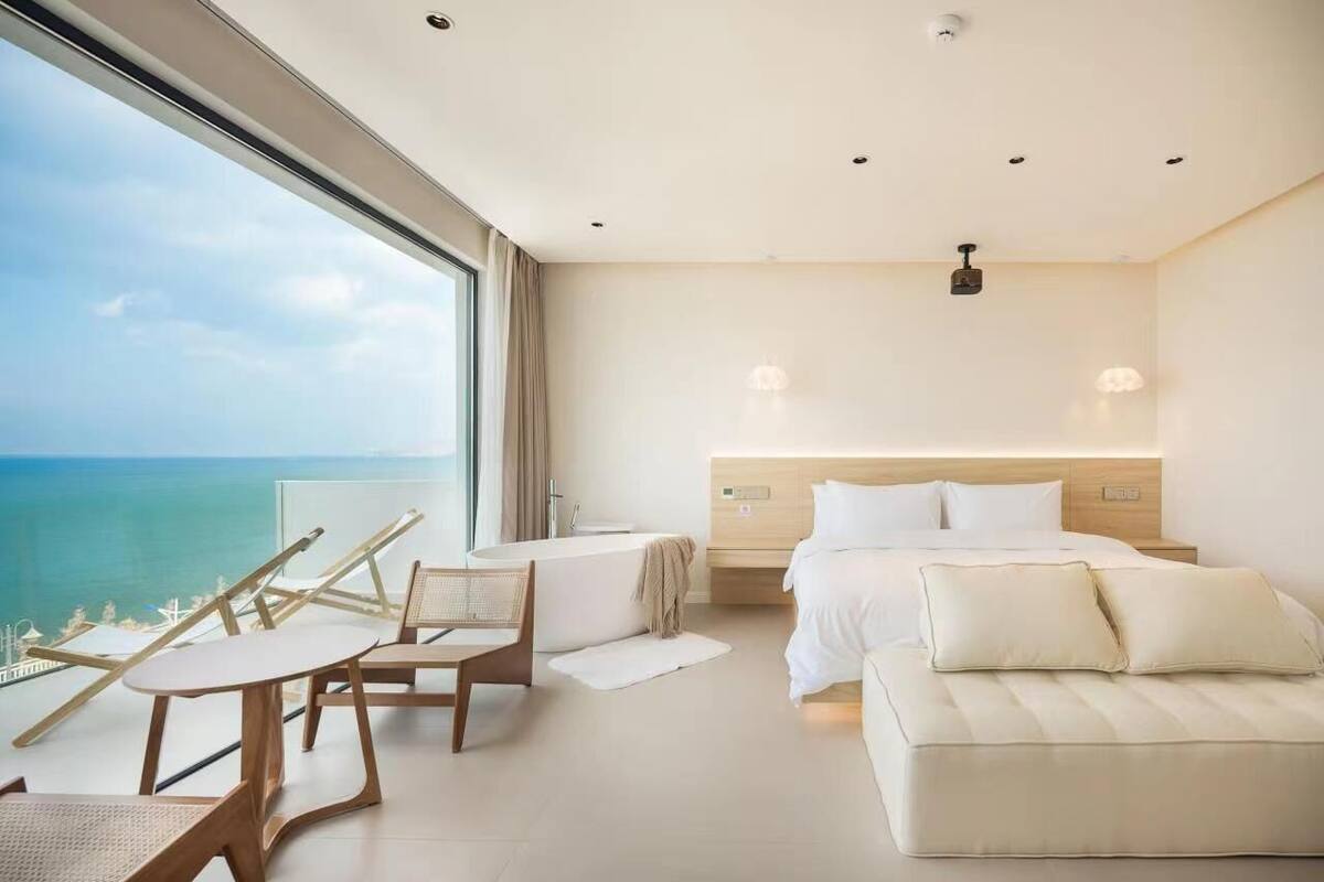 A modern hotel room with a large window overlooking the sea, a bed, a sitting area with a table and chairs, and a bathtub.