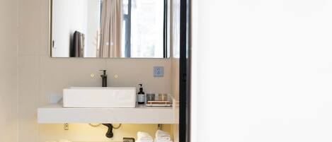 Une salle de bain avec un lavabo blanc, un miroir et une étagère où sont posés des serviettes et des articles de toilette.