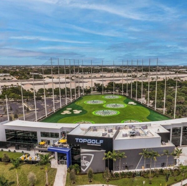 A modern golf course with a TopGolf sign, surrounded by a parking lot and a clear sky.