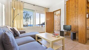Una sala de estar moderna con un sofá, una mesa de centro y una televisión. Hay un ventanal con cortinas, una puerta de madera y vista al mar.