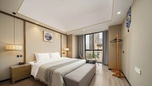 Habitación de hotel moderna con una cama grande, lámparas de noche, una mesita con una silla, perchero y vistas de la ciudad a través de amplios ventanales.