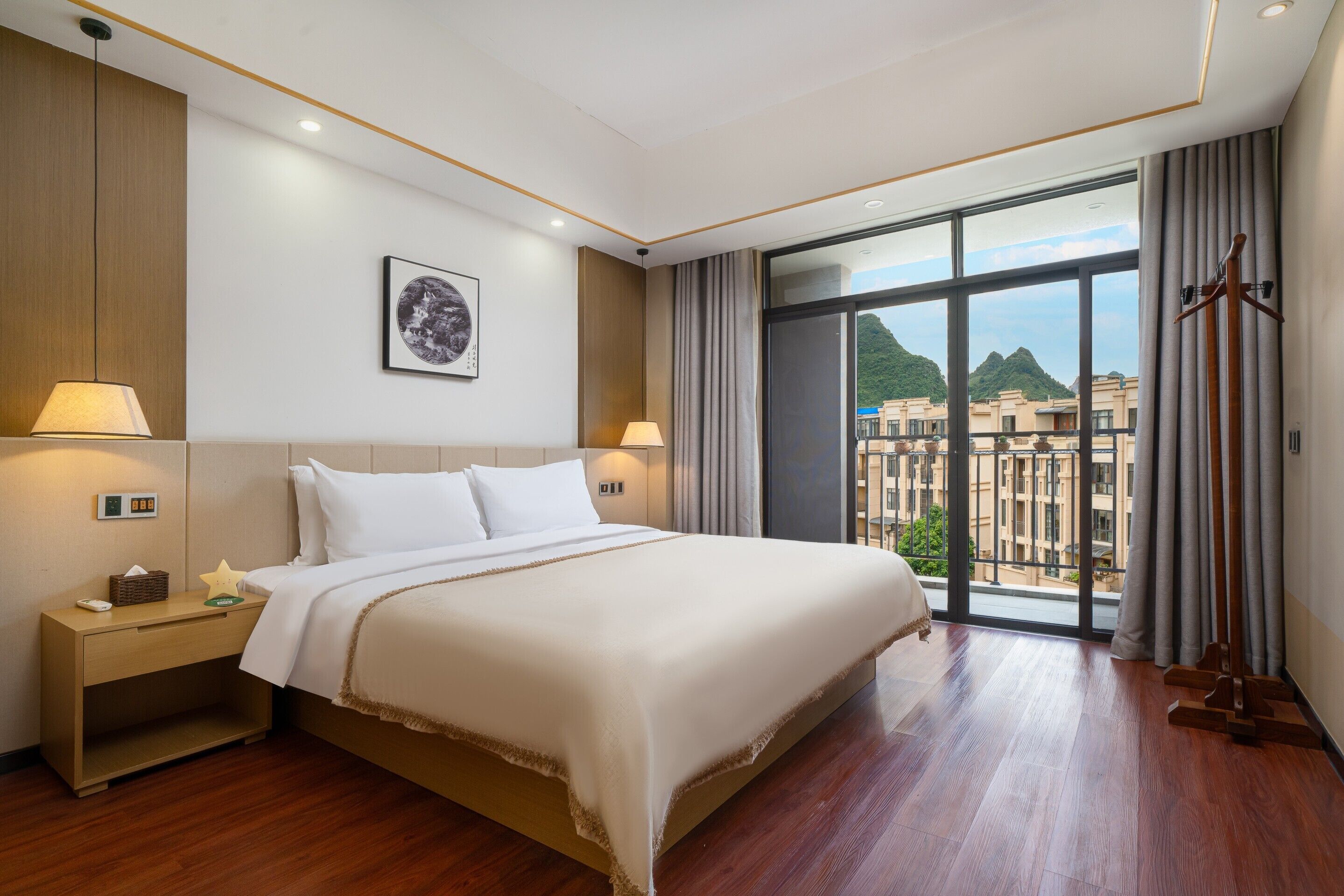 A hotel room with a large bed, bedside lamps, a nightstand, a window with a view of buildings, and a coat rack.