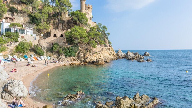 Una zona costera con un castillo en un acantilado, aguas cristalinas y una playa arenosa con sombrillas.
