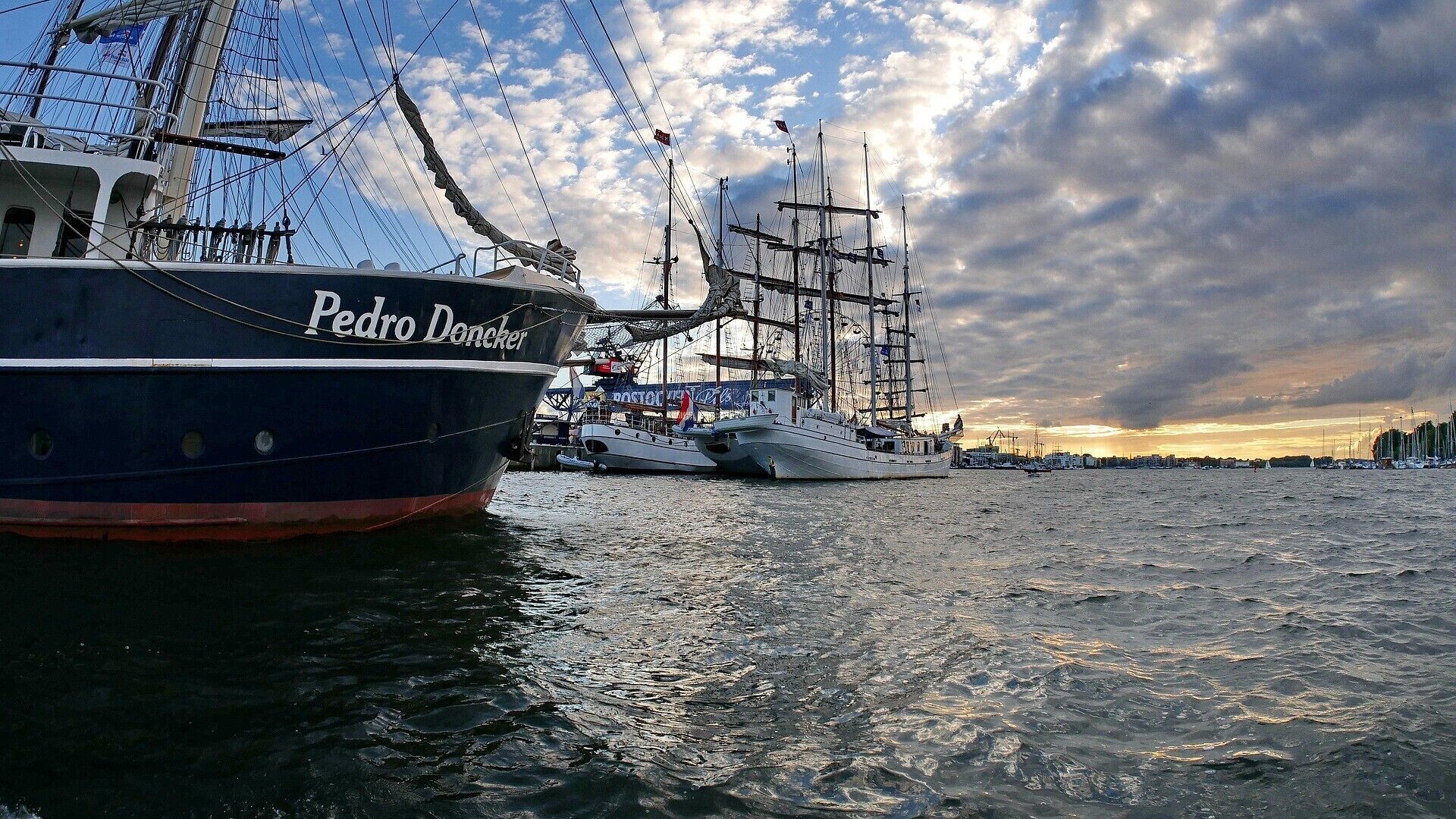 A large ship named 'Pedro Doncker' docked at a harbor with other vessels, under a cloudy sky.