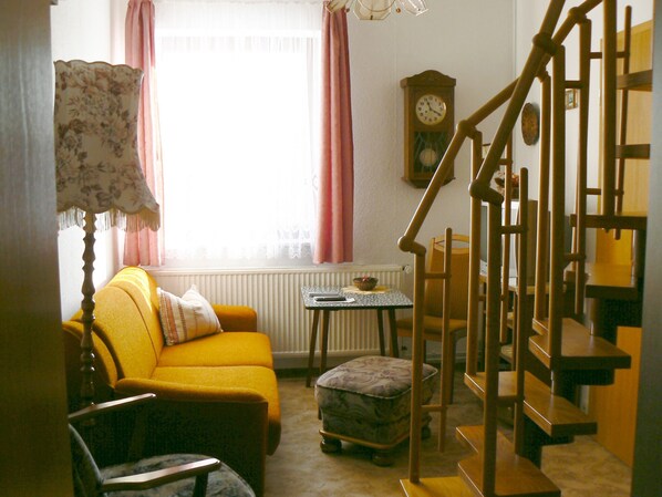 A room with a yellow sofa, a wooden chair, a small table with a bowl, a clock on the wall, and a wooden staircase.