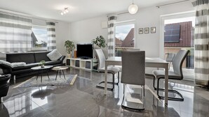 A modern living room with a black leather sofa, a flat-screen TV, a dining table with chairs, and a large window with curtains. - Apartment 'Am Otterbach' with Balcony and Wi-Fi (Jockgrim)