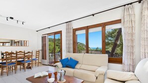 A spacious living room with a sofa, coffee table, and dining area. Large windows offer a view of the outdoors.
