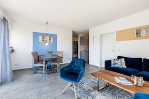 A modern living room with a blue velvet armchair, a wooden coffee table, a blue sofa, and a dining area with a table and chairs.