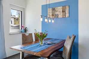 A dining area with a wooden table, chairs, a vase with flowers, a blue wall, and a hanging light fixture.