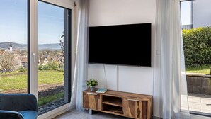 A modern living room with a large TV mounted on the wall, a wooden console table with a plant, and a view of a cityscape through floor-to-ceiling windows. - Apartment 'Untersee' with Lake View, Private Terrace and Wi-Fi (Stockach)