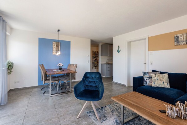 Una sala de estar moderna con un sillón azul, una mesa de centro de madera, un sofá azul oscuro y un comedor con una mesa y sillas de madera.