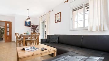 Una sala de estar moderna con un sofá de cuero negro, una mesa de comedor de madera y sillas. Hay una ventana con cortinas blancas y una lámpara colgante.