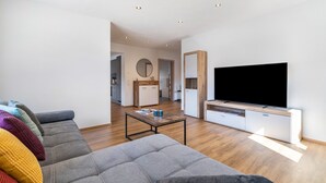 A modern living room with a grey sofa, colorful cushions, a wooden coffee table, a flat-screen TV, and a kitchen in the background. - Apartment 'Winder 1' with Mountain View and Wi-Fi (Lustenau)