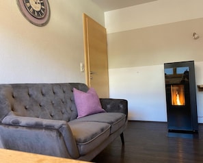 A living room with a grey sofa, a purple pillow, a wall clock, a wooden door, and a fireplace. - Apartment 'Mario' with Mountain View, Private Terrace and Wi-Fi (Hippach)