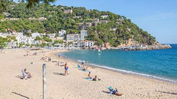 Ein Strand mit Menschen, die sonnenbaden und schwimmen, eine felsige Küste und Gebäude an einem Hügel.