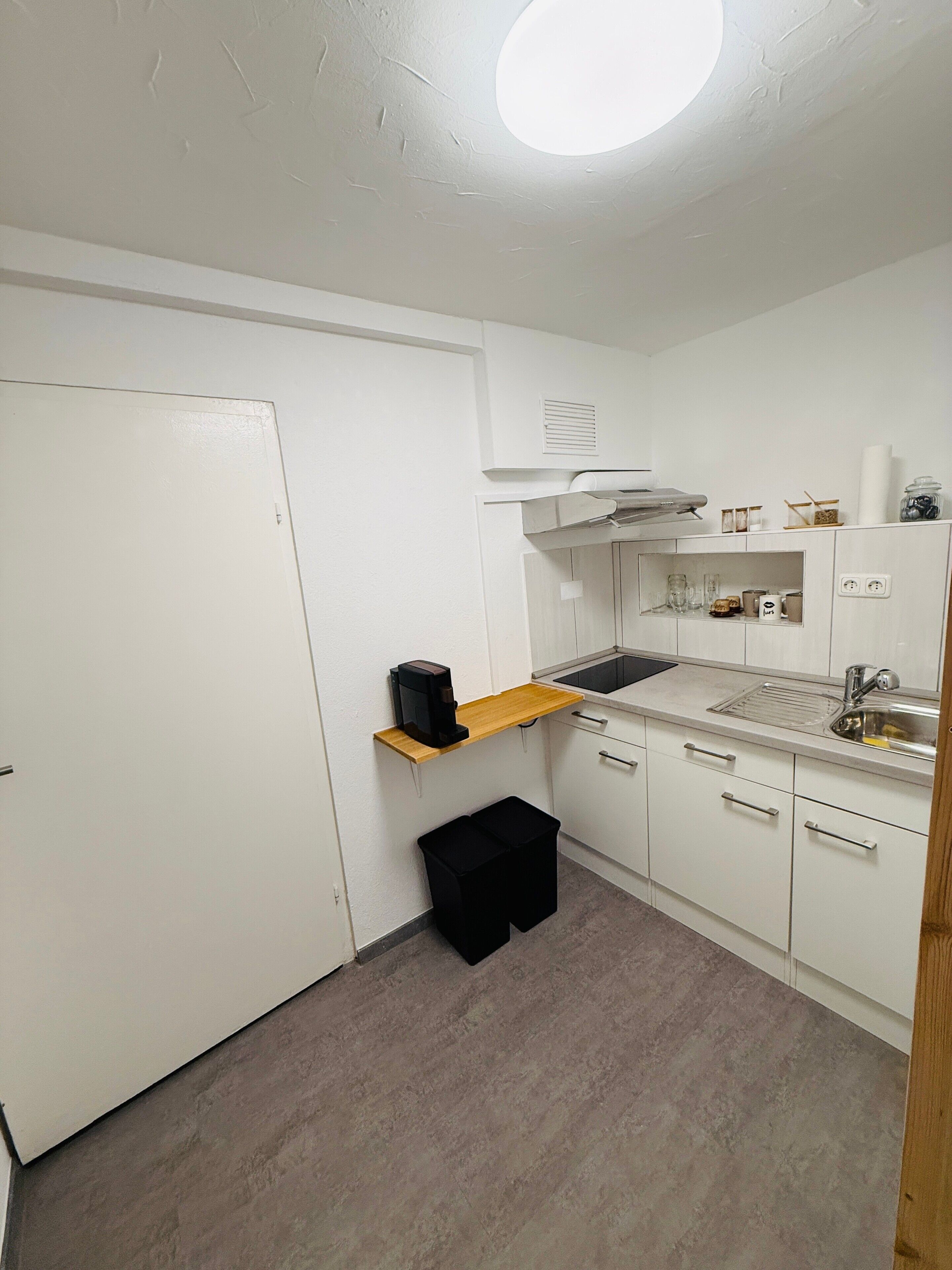 A compact kitchen with white cabinets, a wooden shelf, a black trash bin, a coffee maker, and a sink.
