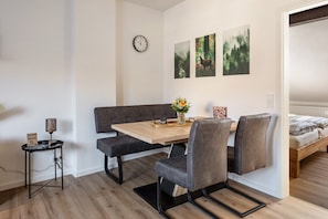 Una habitación moderna con una mesa de comedor de madera, un banco tapizado en gris y dos sillas grises. Un reloj y tres cuadros decoran la pared.