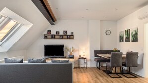 Una sala moderna con sofá, televisión, mesa de comedor y sillas, una mesita auxiliar con lámpara y un reloj en la pared.